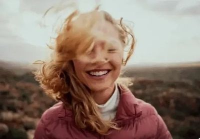 A woman outdoors in a pink jacket, laughing with her hair across her face