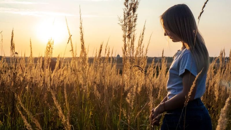 A young woman standing among tall grasses at sunset, her head lowered