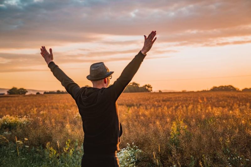 A man in a hat standing in a golden field at sunset, both arms raised to the sky