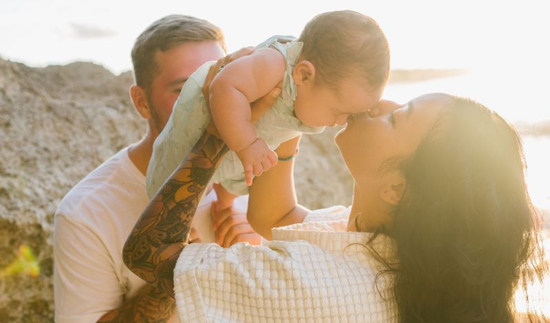 A mother kissing her baby on the nose as the baby’s father lifts her up, sunlight behind them