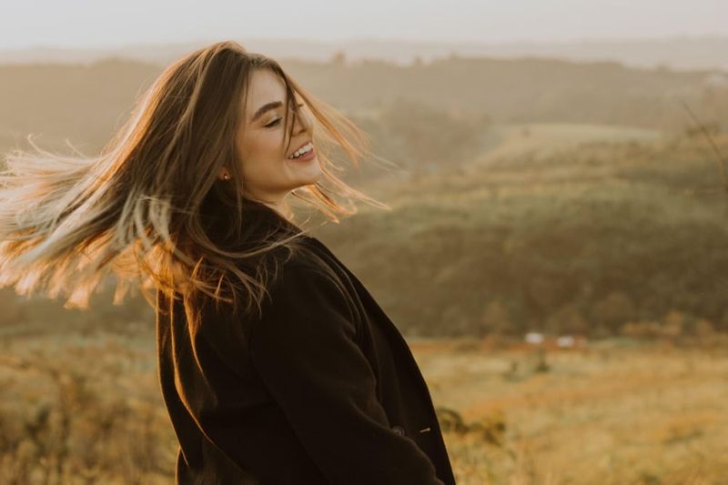 A woman on a hillside at golden hour, her hair blown across her face, laughing with her eyes closed