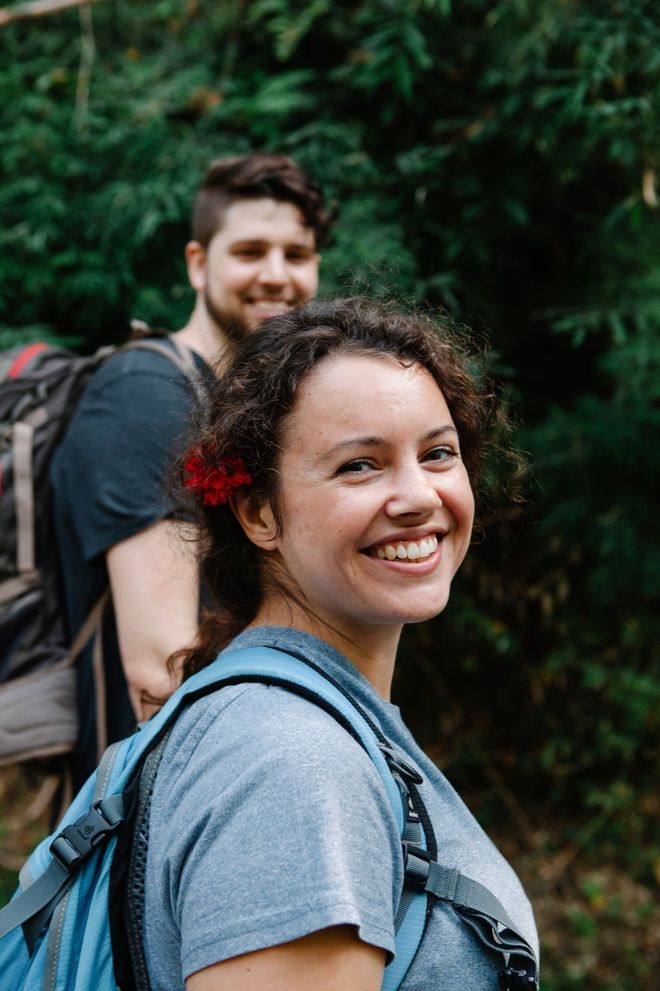 Two people walking a wooded trail with day packs, one turning back to the camera, smiling