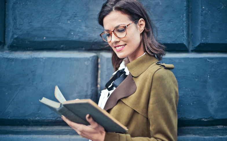 A woman in an olive coat and tortoiseshell glasses reading a book in front of a dark blue wall