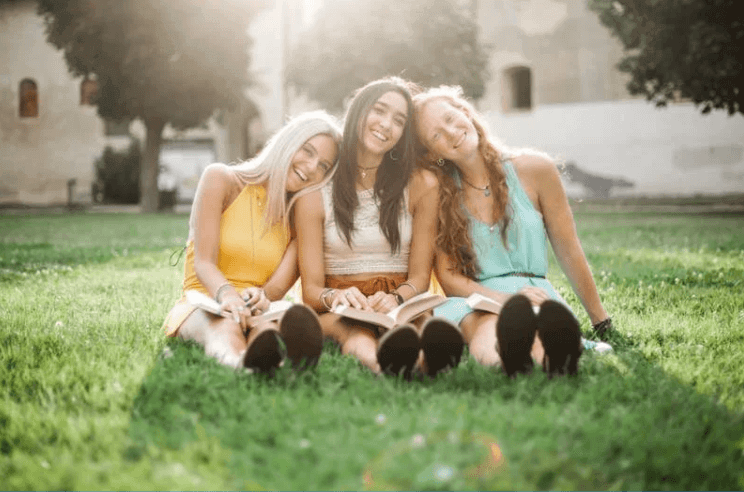 Three teenage girls sitting close together on a lawn with open books, laughing, in low sun