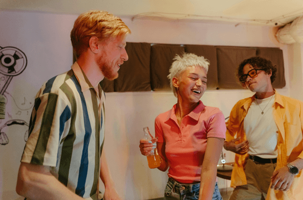 Three young people laughing and talking at a casual indoor gathering, one holding a soda bottle