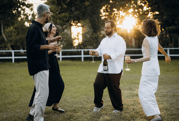 Four adults on a lawn at sunset holding glasses and talking