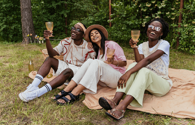 Three friends sitting on a blanket in a wood, laughing, holding glasses