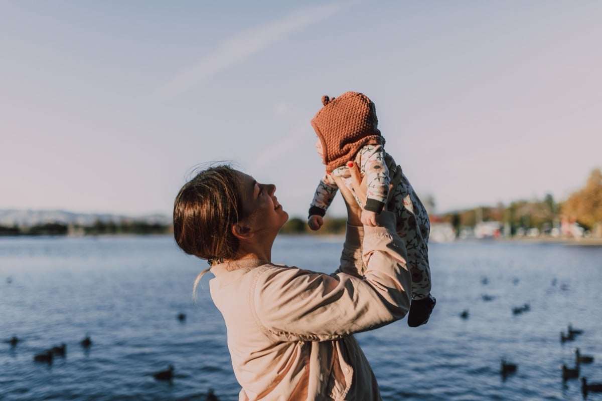 A mother beside a lake lifting her baby, in a knitted hat, up toward the sky