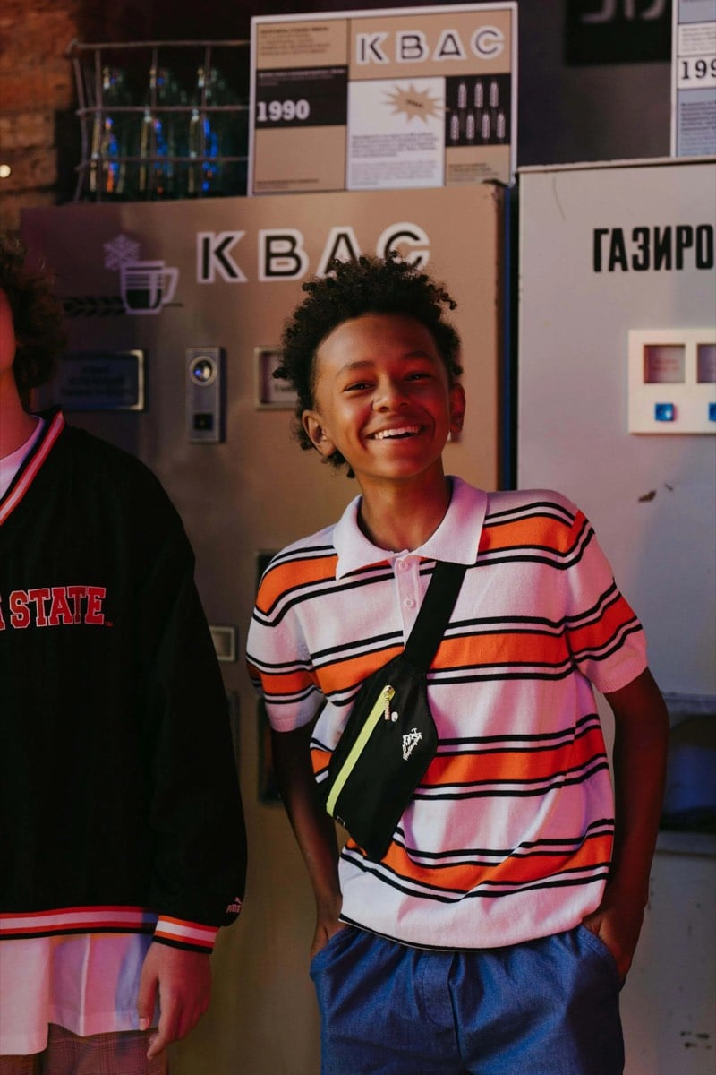 A smiling boy in a striped shirt with a crossbody bag, standing in front of old vending machines