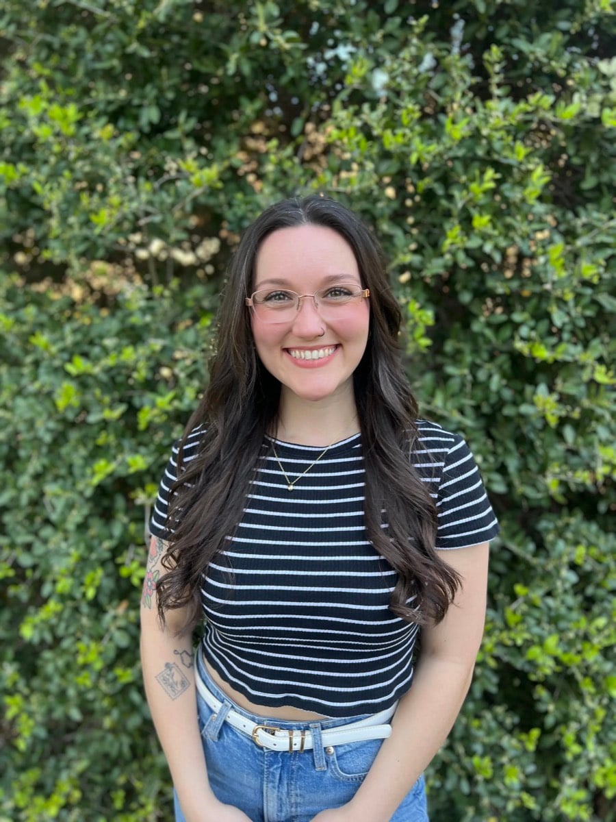 Morganne Fason, in a striped top and glasses in front of a green hedge, smiling