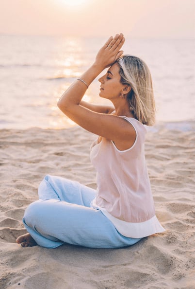A woman sitting cross-legged on a beach at sunrise, hands pressed together against her forehead