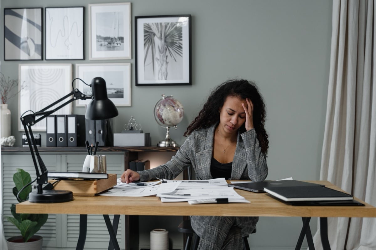 A woman at a desk covered in papers, one hand pressed to her forehead, eyes closed