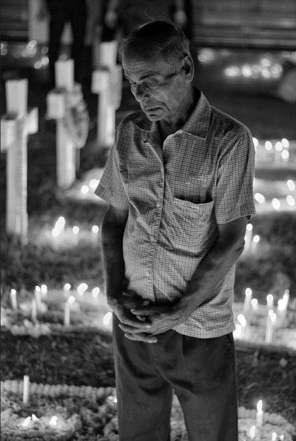 An older man in a checked shirt standing with his head bowed in front of rows of lit memorial candles, in black and white
