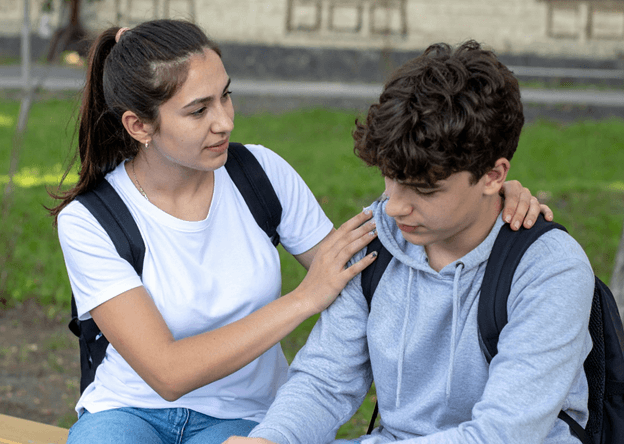A teenage girl with a backpack resting both hands on the shoulders of a teenage boy who is looking down, outdoors on grass