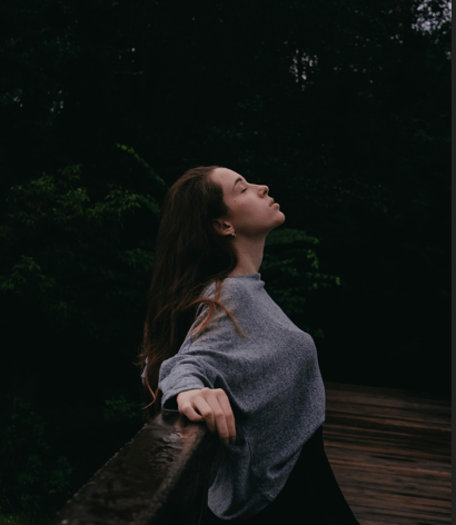A young woman in a grey sweatshirt leaning back against a wooden railing with her eyes closed and her face lifted, trees behind her