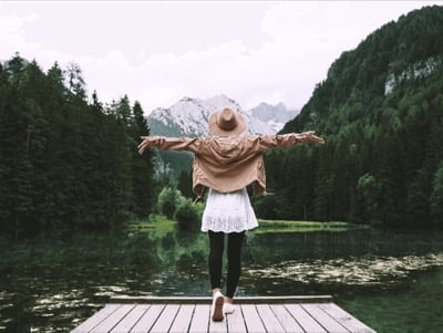 A woman on a wooden jetty with her arms held out, a still lake and mountains ahead of her