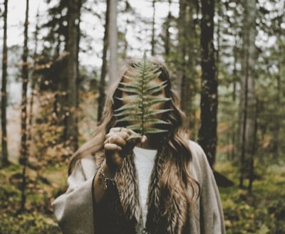 A woman in a wool coat standing in a birch wood, holding a fern frond in front of her face