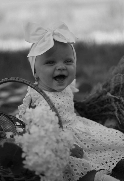 A smiling baby girl in a white bow headscarf sitting outdoors near flowers, in black and white