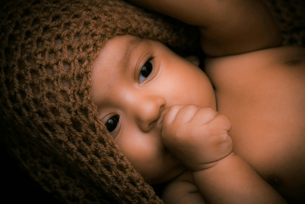 A baby in a brown knitted hat lying down, sucking a thumb