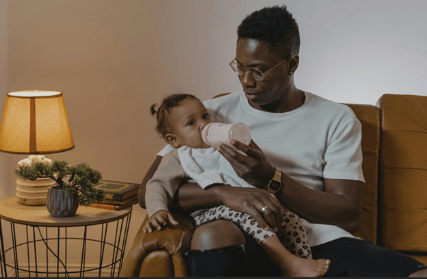 A father in glasses bottle-feeding a baby on his lap in a yellow armchair beside a table lamp