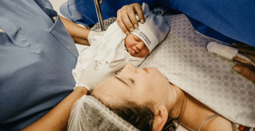 A newborn in a white cap being lifted onto a mother’s chest just after birth, clinicians in blue scrubs beside them