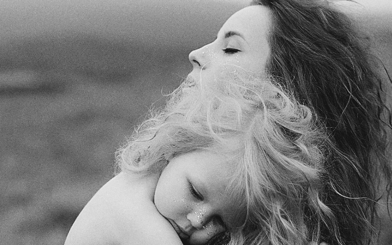 A mother with her eyes closed holding a sleeping toddler against her chest outdoors, in black and white