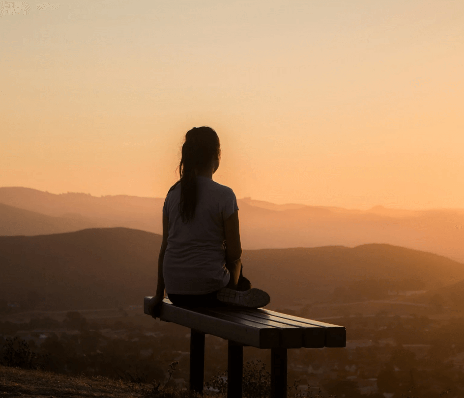 A woman sitting on a bench looking out over hills at sunset