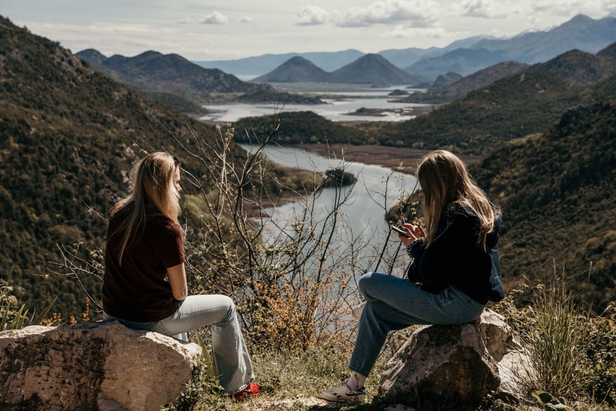 Two women sitting on rocks above a winding river valley, talking, mountains behind them