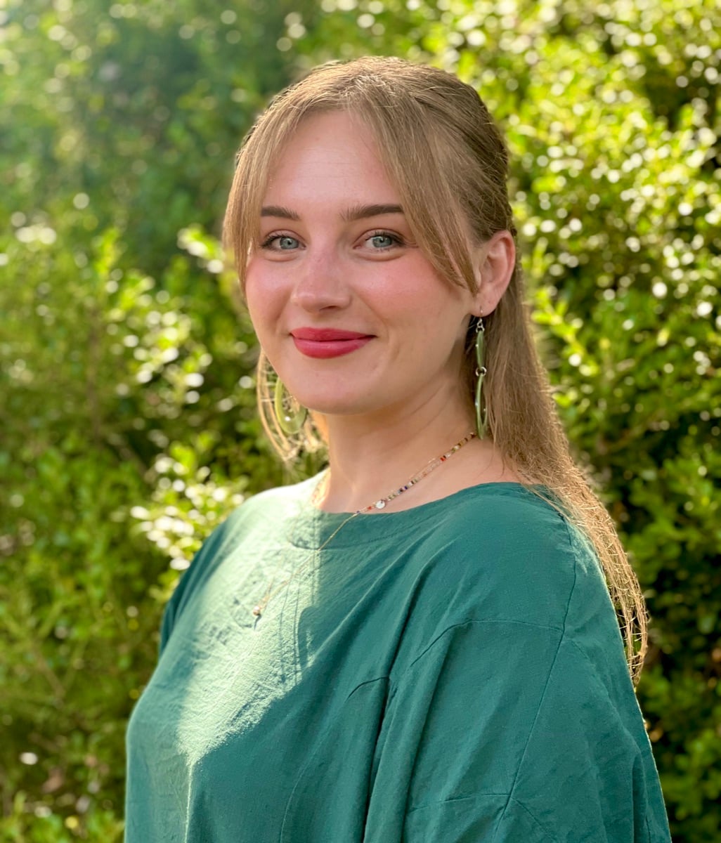 Claire Salander, outdoors in a green top against a flowering hedge, smiling toward the camera
