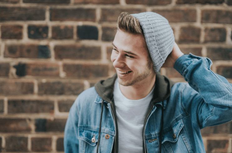 A young man in a denim jacket and gray beanie leaning against a brick wall, laughing and looking off to one side