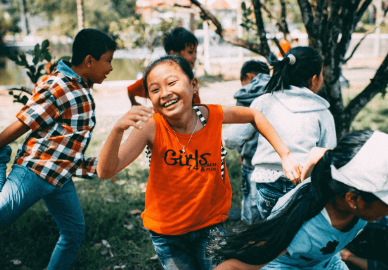 Seven or eight primary-school-age children running and laughing in a park, one girl in an orange vest in sharp focus