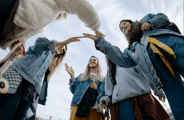 Five teenagers in denim jackets standing in a circle, photographed from below against an overcast sky, reaching their hands toward the center