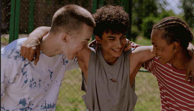 Three teenage boys outdoors in front of a green chain-link fence with their arms around each other’s shoulders, heads bowed together, laughing