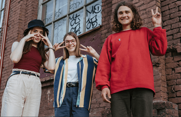 Three teenagers standing against a graffitied brick wall, two making peace signs over their eyes