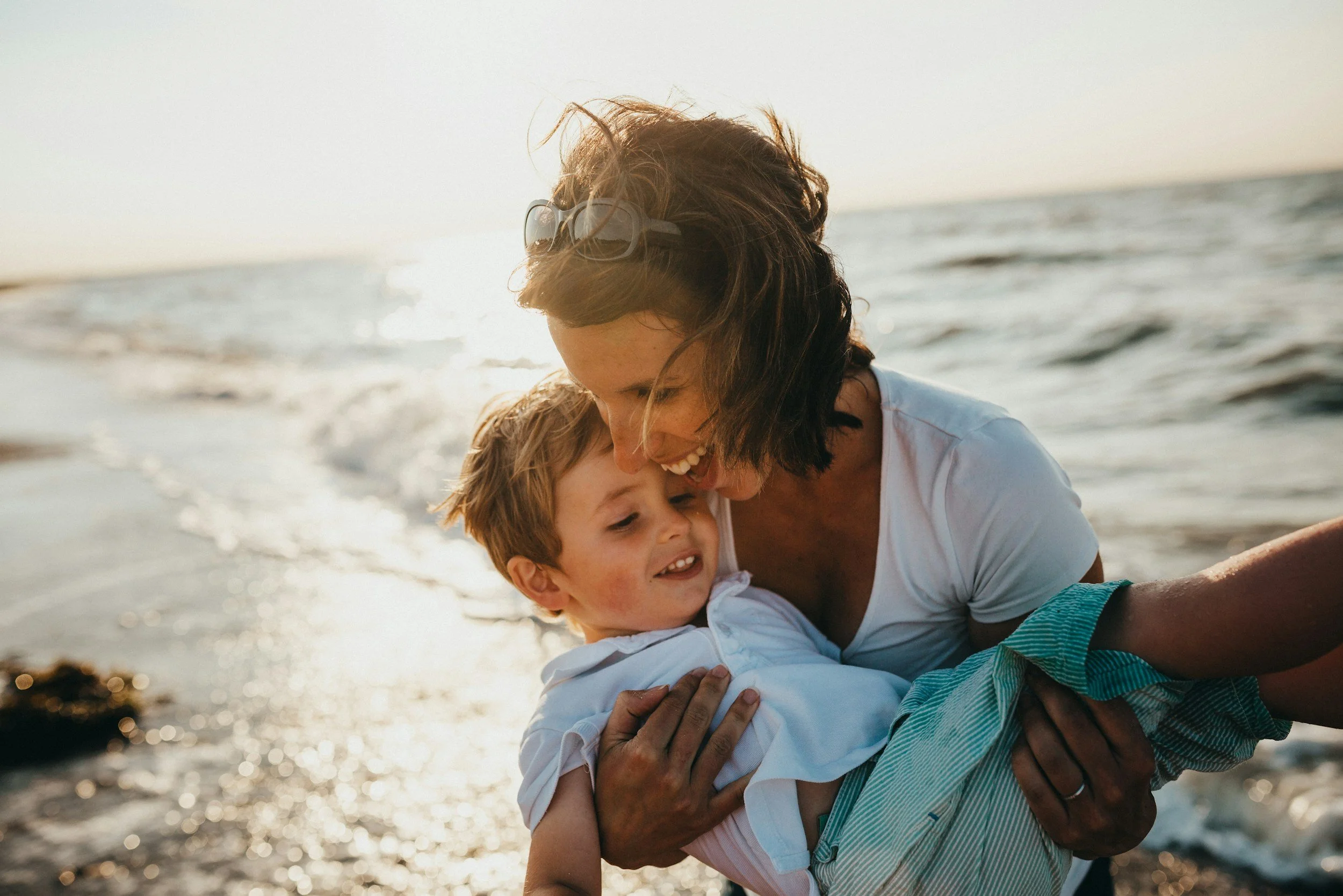 A woman on a beach at sunset lifting a laughing young boy in her arms, waves behind them