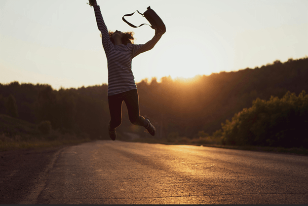 A person jumping in the air on an empty road at sunset, arms raised, boots in one hand