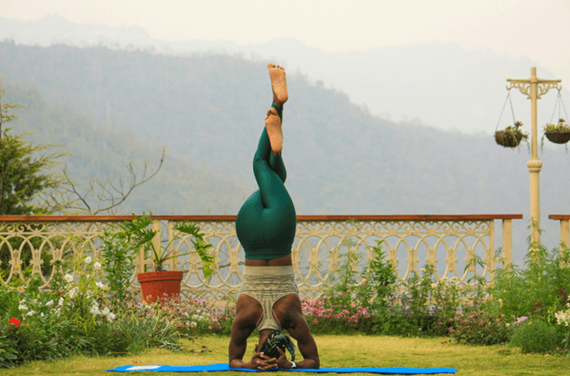 A person in a green outfit holding a supported headstand on a mat on a lawn