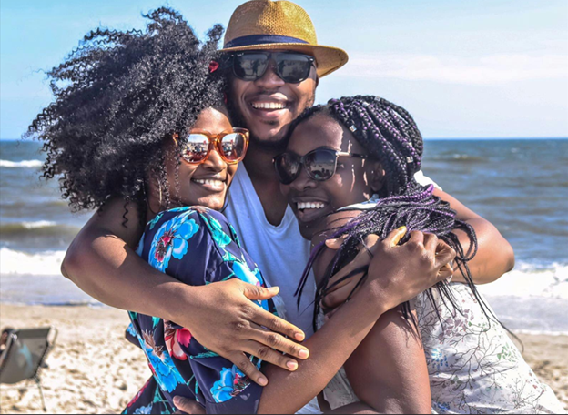 Three friends in sunglasses standing together on a beach with their arms around each other, laughing