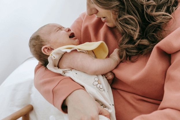 A woman in a rust sweatshirt holding a crying newborn on her lap