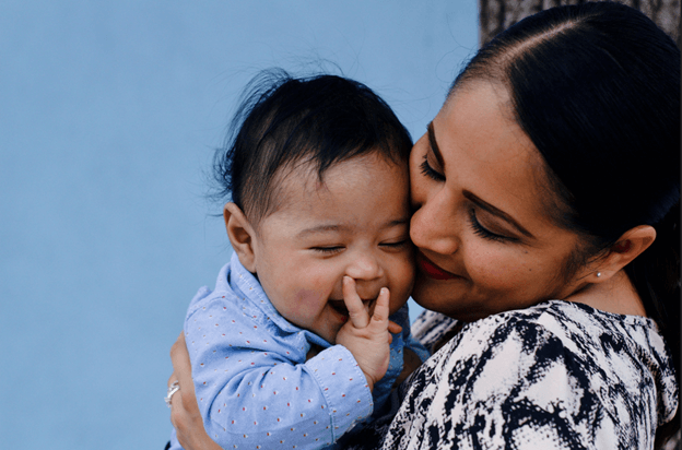 A woman holding a laughing baby cheek to cheek in front of a blue wall