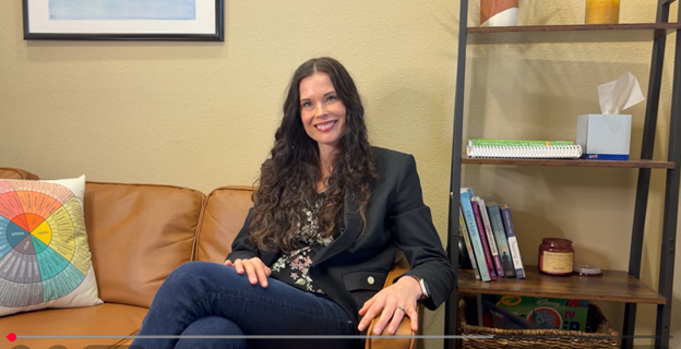 A therapist in a black blazer sitting on a tan leather sofa in a warmly lit office, smiling toward the camera, a bookshelf and a framed print behind her