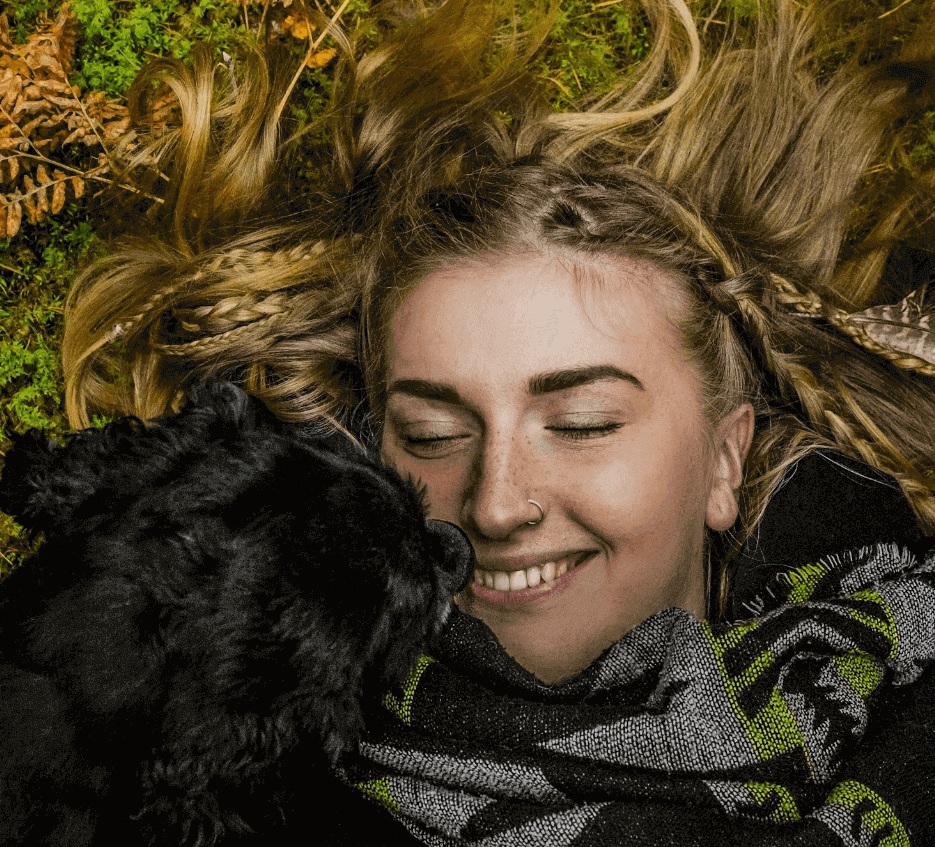 A woman lying back in moss and bracken with her eyes closed, a black dog’s nose against her cheek