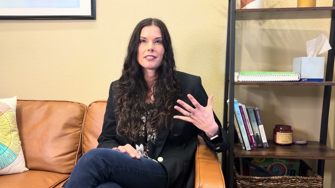 A therapist seated on a sofa in a warmly lit office, speaking to camera