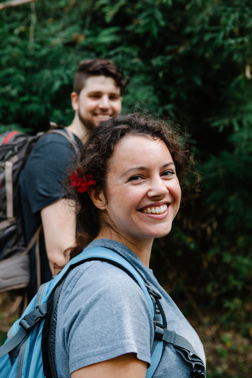 Two people walking a wooded trail with day packs, one turning back to the camera, smiling